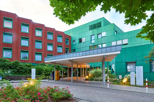 Exterior view of a modern medical specialist clinic. A modern multi-story medical facility with a distinctive green and red facade, featuring a covered entrance area and landscaped gardens with flowers.